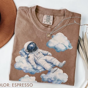 Peut inclure: T-shirt couleur expresso avec un motif d'astronaute allong&eacute; sur un nuage, avec des nuages bleus et blancs &agrave; l'aquarelle. Le t-shirt a un col rond et une petite &eacute;tiquette au niveau de l'encolure. Un collier argent&eacute; avec un pendentif est drap&eacute; sur le t-shirt.