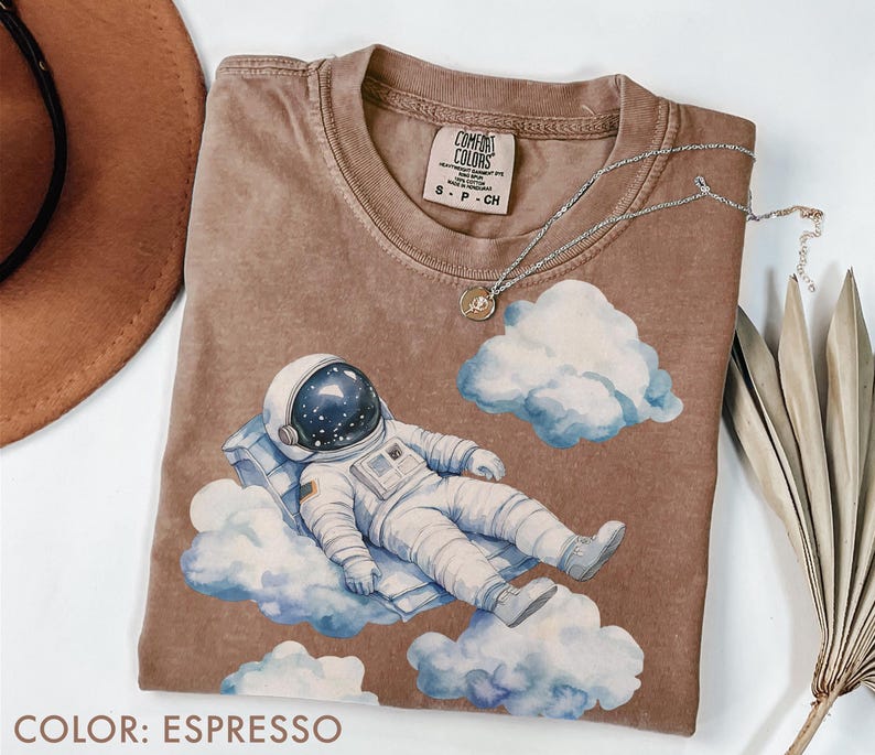 Peut inclure: T-shirt couleur expresso avec un motif d'astronaute allong&eacute; sur un nuage, avec des nuages bleus et blancs &agrave; l'aquarelle. Le t-shirt a un col rond et une petite &eacute;tiquette au niveau de l'encolure. Un collier argent&eacute; avec un pendentif est drap&eacute; sur le t-shirt.