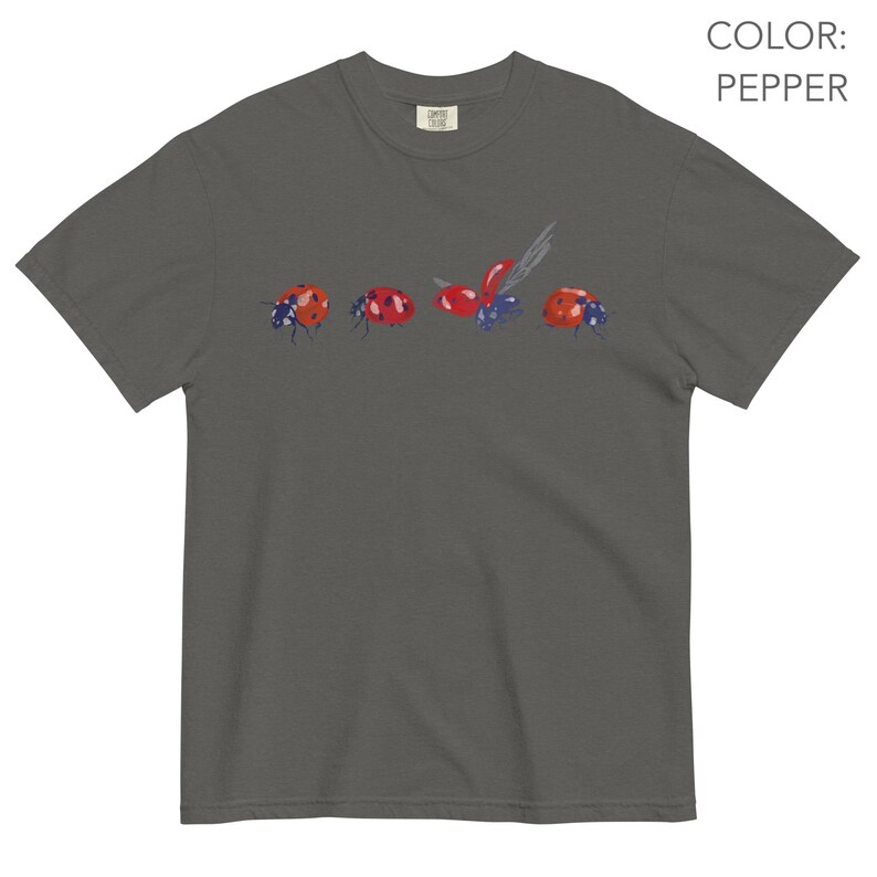May include: Dark grey t-shirt with a watercolour illustration of ladybirds. The ladybirds are red with black spots and are set against a dark grey background. The shirt has a crew neck and short sleeves. The colour is 'pepper'.