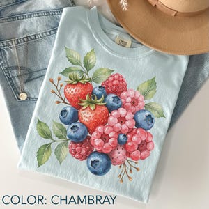 May include: Pale blue t-shirt featuring a watercolour design of strawberries, raspberries, and blueberries. The shirt is labelled "Comfort Colors" and the colour is "Chambray". The shirt is folded on top of blue jeans.