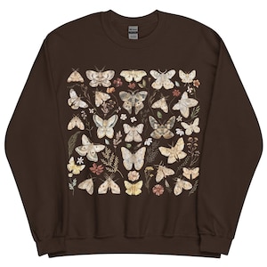 May include: A dark brown crewneck sweatshirt featuring a central graphic print of various moths and small wildflowers in muted earth tones. The design displays multiple moth species with intricate wing patterns arranged among botanical elements.