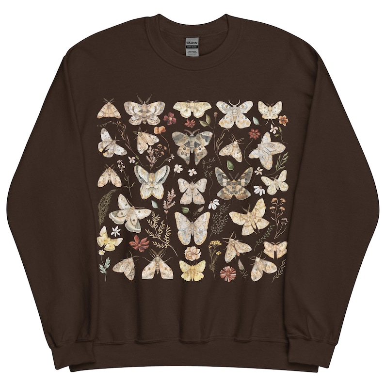 May include: A dark brown crewneck sweatshirt featuring a central graphic print of various moths and small wildflowers in muted earth tones. The design displays multiple moth species with intricate wing patterns arranged among botanical elements.