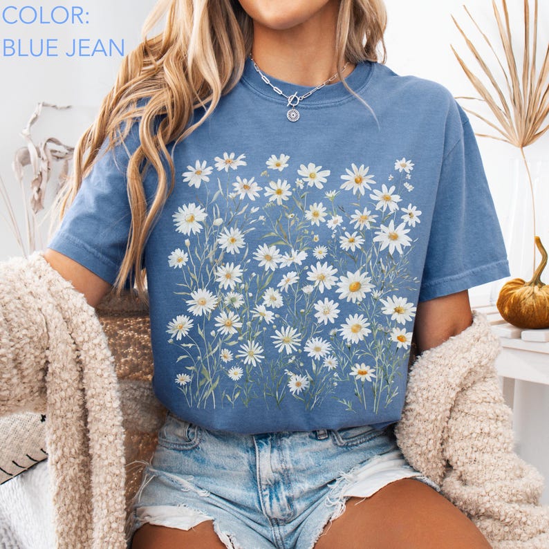 May include: Blue jean colored t-shirt with a daisy floral design. The shirt features a crew neck and short sleeves. The text "COLOR: BLUE JEAN" is visible in the upper left corner. The shirt is paired with denim shorts.