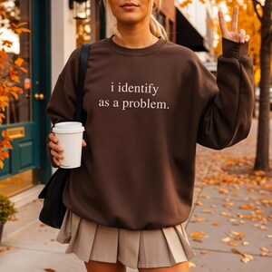 Könnte beinhalten: Ein dunkelbraunes Sweatshirt mit dem weißen Text „i identify as a problem“. Das Sweatshirt wird mit einem beigefarbenen Faltenrock getragen. Eine Person hält eine weiße Kaffeetasse und zeigt das Friedenszeichen.
