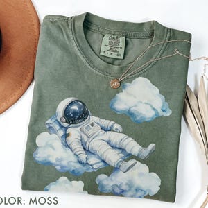 Peut inclure: T-shirt vert mousse avec une illustration &agrave; l'aquarelle d'un astronaute se relaxant sur des nuages. L'astronaute porte une combinaison spatiale et un casque blancs. Le t-shirt a un col rond et des manches courtes. Le texte "COLOR: MOSS" est en bas.
