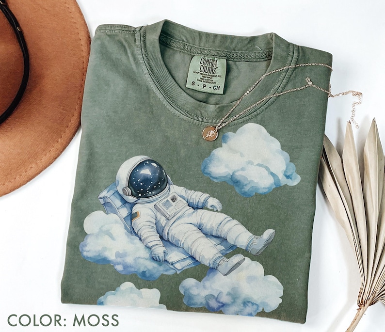 Peut inclure: T-shirt vert mousse avec une illustration &agrave; l'aquarelle d'un astronaute se relaxant sur des nuages. L'astronaute porte une combinaison spatiale et un casque blancs. Le t-shirt a un col rond et des manches courtes. Le texte "COLOR: MOSS" est en bas.