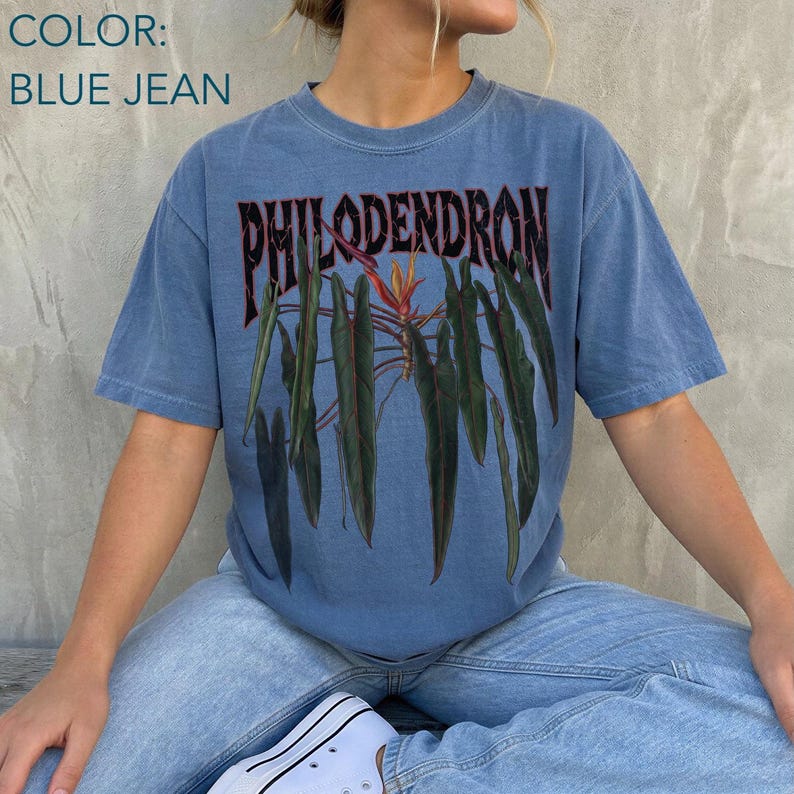 May include: A blue jean colored short-sleeve t-shirt featuring a graphic print of dark green philodendron leaves and a central orange flower. The word PHILODENDRON is printed in a bold, dark, stylized font above the botanical illustration.