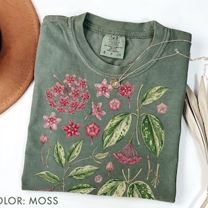 May include: A moss-colored t-shirt featuring a botanical illustration of pink flowers and green leaves. The shirt has a crew neck and short sleeves. A gold necklace with a pendant is draped across the shirt. The text "COLOR: MOSS" is at the bottom.