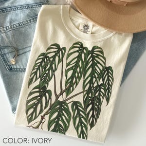 May include: An ivory-coloured crew neck t-shirt featuring a large, detailed graphic of a green monstera plant with characteristic fenestrated leaves. The shirt has a Comfort Colors brand label printed on the inside of the collar area.