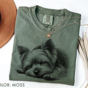 Puede incluir: Una camiseta verde musgo con una ilustraci&oacute;n detallada de un Yorkshire Terrier durmiendo. La camiseta tiene cuello redondo y mangas cortas. Un delicado collar de oro con un peque&ntilde;o colgante descansa sobre la camiseta. El texto "COLOR: MOSS" est&aacute; en la parte inferior.