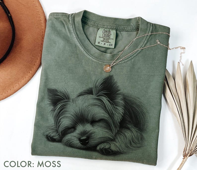 Puede incluir: Una camiseta verde musgo con una ilustraci&oacute;n detallada de un Yorkshire Terrier durmiendo. La camiseta tiene cuello redondo y mangas cortas. Un delicado collar de oro con un peque&ntilde;o colgante descansa sobre la camiseta. El texto "COLOR: MOSS" est&aacute; en la parte inferior.