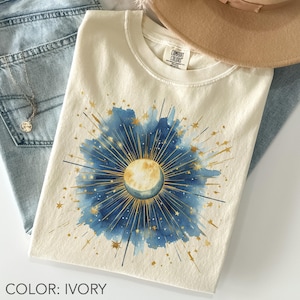 May include: Ivory-colored t-shirt featuring a celestial design with a watercolor effect. The design includes a crescent moon, stars, and sunbursts in shades of blue and gold. The shirt is folded on top of blue jeans.