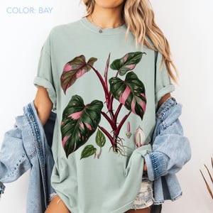Peut inclure: T-shirt vert clair avec une illustration botanique d'une plante. La plante a de grandes feuilles vertes avec des marques roses et une tige rouge fonc&eacute;. Le texte "COLOR: BAY" est en haut &agrave; gauche.