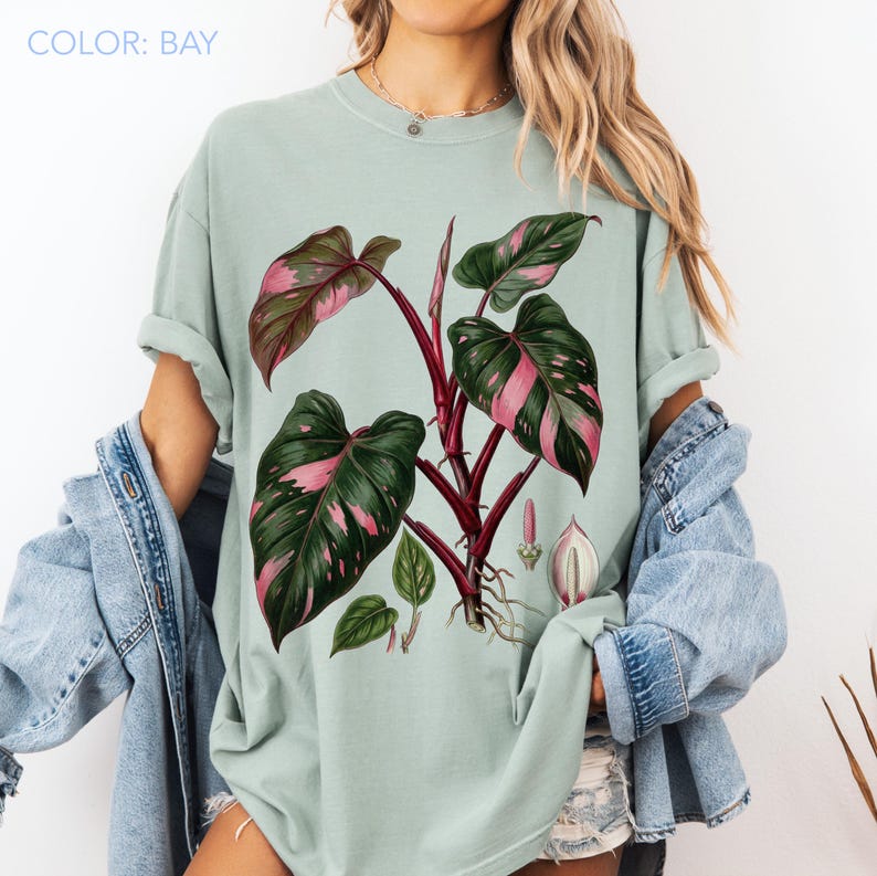Peut inclure: T-shirt vert clair avec une illustration botanique d'une plante. La plante a de grandes feuilles vertes avec des marques roses et une tige rouge fonc&eacute;. Le texte "COLOR: BAY" est en haut &agrave; gauche.