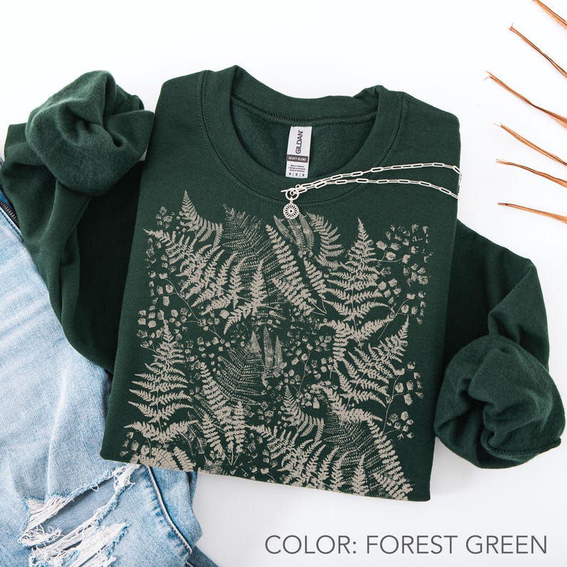 Puede incluir: Sudadera verde bosque con dise&ntilde;o de helechos y collar plateado. La sudadera est&aacute; doblada, con unos vaqueros azules y una sudadera verde a juego visibles. El texto "COLOR: FOREST GREEN" est&aacute; en la parte inferior de la imagen.