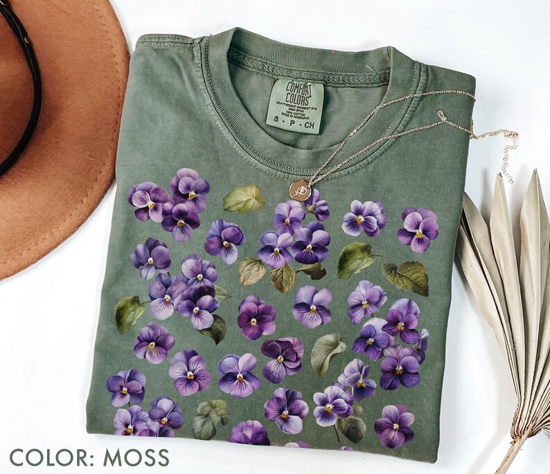 May include: Moss green t-shirt with a floral design of purple pansies and green leaves. The shirt has a crew neck and short sleeves. A gold necklace with a pendant is draped across the shirt. The text "COLOR: MOSS" is at the bottom.