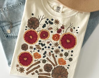 May include: Ivory-colored t-shirt with a botanical design featuring red citrus slices, brown pinecones, and cinnamon sticks. The shirt is laid on top of blue jeans. The text "COLOR: IVORY" is at the bottom of the image.