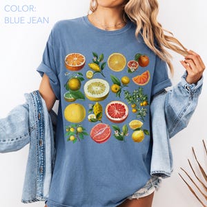May include: A blue jean colored t-shirt featuring a graphic print of various citrus fruits including oranges, grapefruits, and lemons with green leaves and white blossoms. The design displays whole and sliced fruits in shades of yellow, orange, and red.