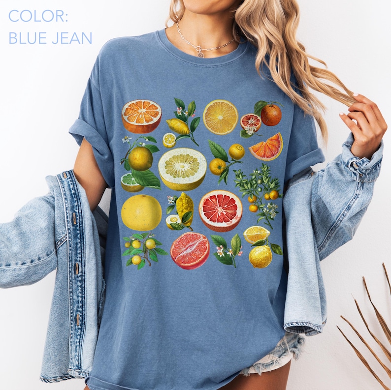 May include: A blue jean colored t-shirt featuring a graphic print of various citrus fruits including oranges, grapefruits, and lemons with green leaves and white blossoms. The design displays whole and sliced fruits in shades of yellow, orange, and red.