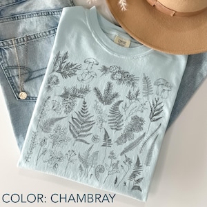 May include: A light blue t-shirt with a botanical illustration of ferns, mushrooms, and pinecones. The shirt is folded on top of a pair of blue jeans. The text "COLOR: CHAMBRAY" is at the bottom of the image.