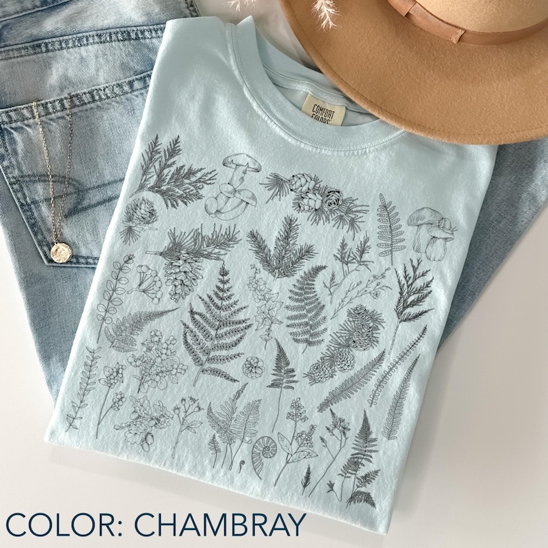 May include: A light blue t-shirt with a botanical illustration of ferns, mushrooms, and pinecones. The shirt is folded on top of a pair of blue jeans. The text "COLOR: CHAMBRAY" is at the bottom of the image.