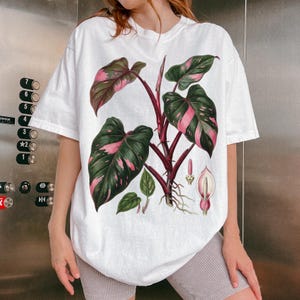 Peut inclure: T-shirt blanc avec une illustration botanique d'une plante philodendron princesse rose. Le motif montre les feuilles, les tiges et les racines de la plante. Les feuilles sont vertes avec des panachures roses.