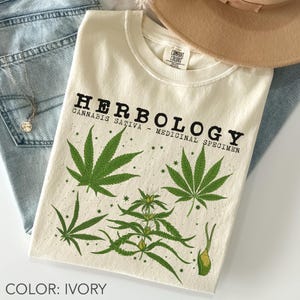 Peut inclure: T-shirt ivoire avec le mot "HERBOLOGY" imprimé en noir, ainsi que "CANNABIS SATIVA - MEDICINAL SPECIMEN". Le t-shirt présente des illustrations de feuilles de cannabis vertes. Un chapeau et un jean sont en arrière-plan.