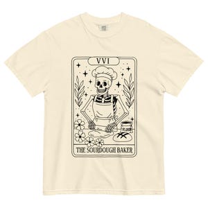 May include: Cream-colored t-shirt featuring a black and white illustration of a skeleton baker wearing a chef's hat and apron. The design includes the text "THE SOURDOUGH BAKER" and the Roman numeral "VVI". The shirt has a classic crew neck.