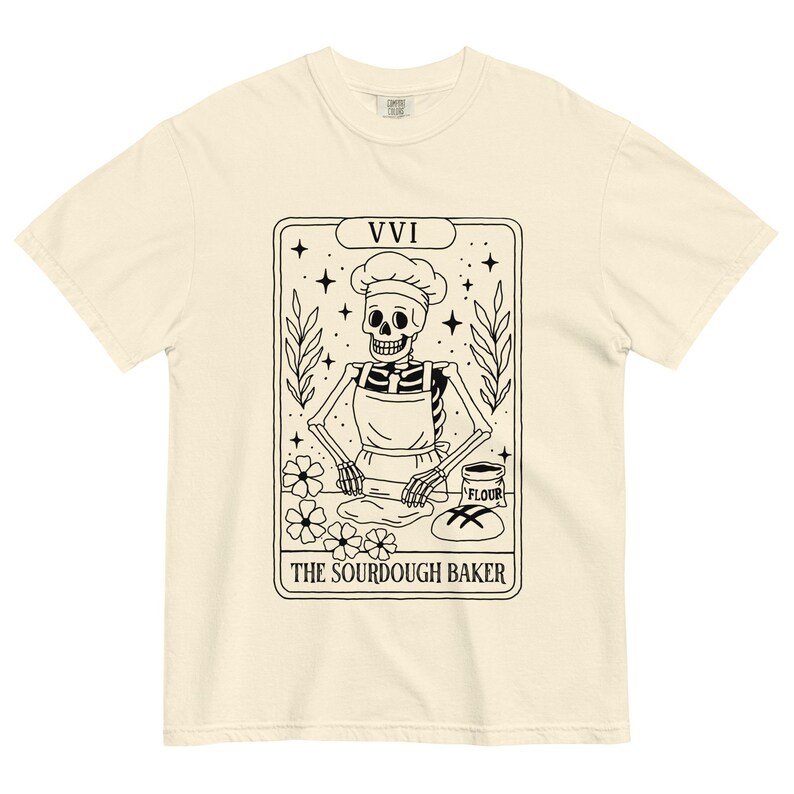 May include: Cream-colored t-shirt featuring a black and white illustration of a skeleton baker wearing a chef's hat and apron. The design includes the text "THE SOURDOUGH BAKER" and the Roman numeral "VVI". The shirt has a classic crew neck.