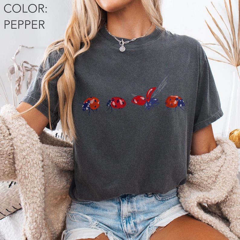 May include: A dark grey t-shirt with a ladybug design. The shirt features four ladybug illustrations in red, orange, and blue. The text "COLOR: PEPPER" is in the upper left corner. The shirt is worn by a person.