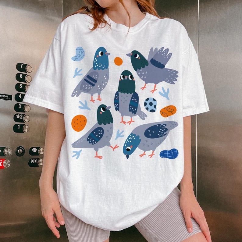 May include: White t-shirt featuring a whimsical illustration of several pigeons in shades of blue, gray, and teal, with orange and blue accent shapes. The design is printed on a soft, casual tee.