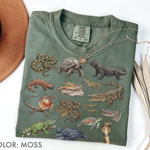 Könnte beinhalten: Ein moosgrünes T-Shirt mit einem detaillierten Grafikdruck verschiedener Reptilien und Amphibien, darunter Schlangen, Schildkröten, ein Krokodil, ein Leguan und eine Eidechse. Das Shirt hat einen Rundhalsausschnitt und ein Comfort Colors Etikett.