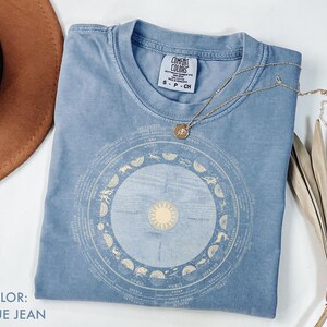 Pode incluir: Uma camiseta azul jeans com um design astrol&oacute;gico circular em bege claro. A camisa tem gola redonda e mangas curtas. Um colar de ouro com um pingente est&aacute; drapeado sobre a camisa. A camisa est&aacute; dobrada sobre uma superf&iacute;cie branca.