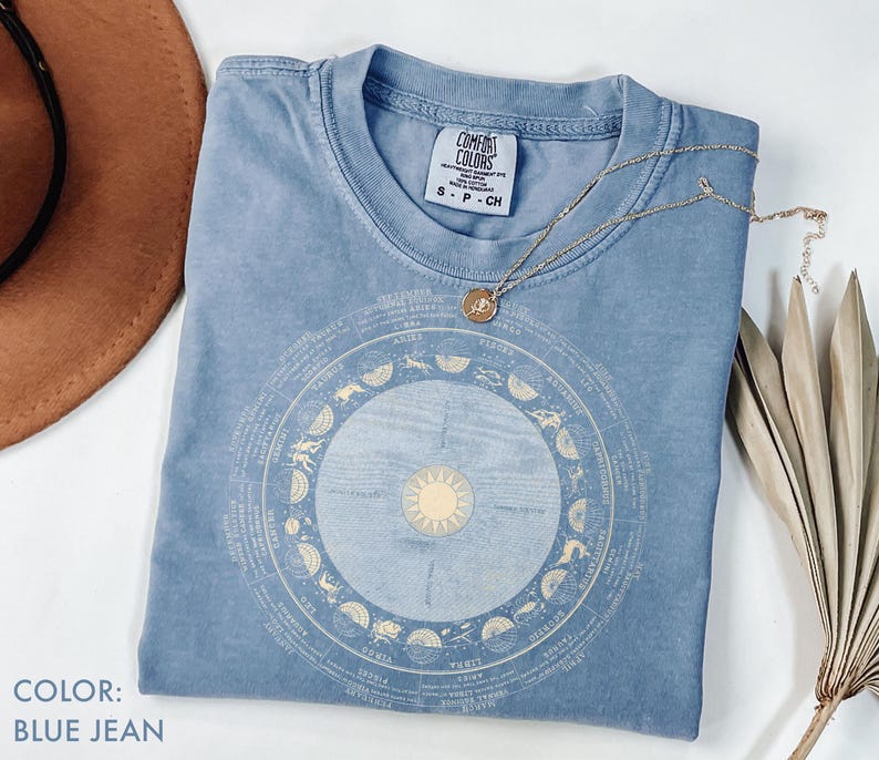 Pode incluir: Uma camiseta azul jeans com um design astrol&oacute;gico circular em bege claro. A camisa tem gola redonda e mangas curtas. Um colar de ouro com um pingente est&aacute; drapeado sobre a camisa. A camisa est&aacute; dobrada sobre uma superf&iacute;cie branca.