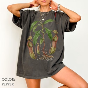 Può includere: T-shirt color pepe con illustrazione botanica di una pianta carnivora. La grafica include foglie verdi, brocche rosso-brunastre e radici esposte. La maglietta ha una vestibilità ampia e maniche corte. Il testo "COLOR: PEPPER" è in basso a sinistra.
