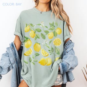 May include: A light green t-shirt featuring a watercolor design of lemons, leaves, and white flowers. The lemons are yellow, and the leaves are green. The text "COLOR: BAY" is in the upper left corner.