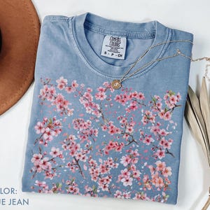 May include: Blue jean colored t-shirt with a floral cherry blossom design. The shirt has a crew neck and short sleeves. A gold necklace with a pendant is draped across the shirt. The text "COLOR: BLUE JEAN" is in the bottom left corner.