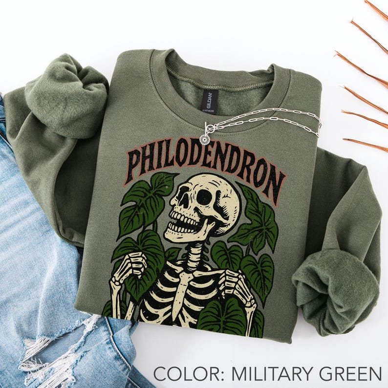 May include: Military green crewneck sweatshirt with a skeleton graphic surrounded by green leaves and the word "PHILODENDRON" in a red and brown font. A silver necklace with a small pendant is draped across the sweatshirt.