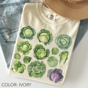 May include: Ivory-coloured t-shirt with a watercolour design of various cabbages and leafy greens. The shirt features different types of cabbage, including green and purple varieties. The text "COLOR: IVORY" is printed at the bottom of the image.