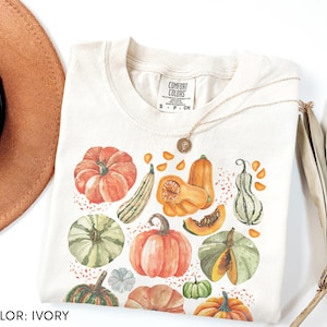 May include: Ivory Comfort Colors t-shirt with a watercolour design of pumpkins and gourds in orange, green, and yellow. The shirt has a crew neck and short sleeves. A gold necklace is draped across the shirt.