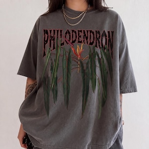 May include: A charcoal gray short-sleeve t-shirt featuring a graphic print of green philodendron leaves and a central orange and yellow flower. The word PHILODENDRON is printed in a bold, dark, distressed font above the botanical illustration.