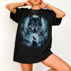 K&ouml;nnte beinhalten: Schwarzes T-Shirt mit detaillierter Grafik von W&ouml;lfen unter Vollmond und Blitzen. Das Design umfasst ein zentrales Wolfsgesicht und drei heulende W&ouml;lfe vor einem Waldhintergrund. Das Shirt ist ein l&auml;ssiger, &uuml;bergro&szlig;er Stil.