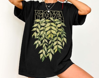 May include: A black short-sleeve t-shirt featuring a graphic print of hanging green leaves and the text HOYA in a bold, distressed font. The design is centered on the chest, showcasing a botanical illustration with varied shades of green.