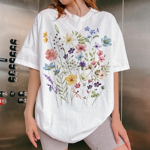 Puede incluir: Camiseta blanca con un dise&ntilde;o floral en acuarela. El dise&ntilde;o incluye una variedad de flores coloridas, incluyendo flores rosas, amarillas, azules y moradas, junto con hojas y tallos verdes. La camiseta tiene mangas cortas y un corte relajado.
