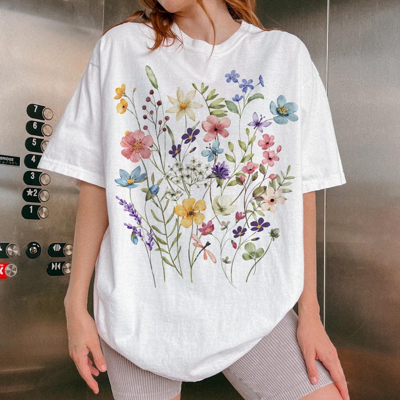 Puede incluir: Camiseta blanca con un dise&ntilde;o floral en acuarela. El dise&ntilde;o incluye una variedad de flores coloridas, incluyendo flores rosas, amarillas, azules y moradas, junto con hojas y tallos verdes. La camiseta tiene mangas cortas y un corte relajado.