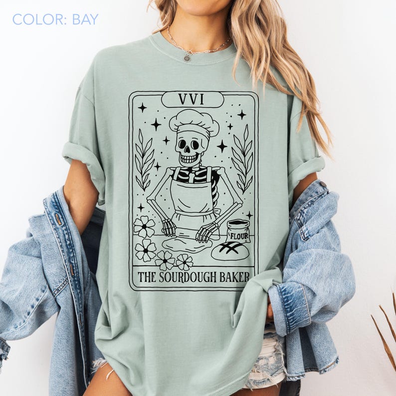 May include: A light green t-shirt featuring a black and white graphic of a skeleton baker wearing a chef's hat and apron, with the text "THE SOURDOUGH BAKER". The design includes stars, flowers, and the Roman numeral "VVI".