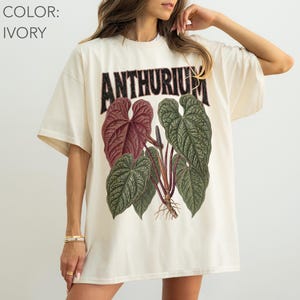 May include: An ivory short-sleeved t-shirt featuring a graphic print of an Anthurium plant with large, textured green and deep red leaves. The word ANTHURIUM is printed in a bold, dark, distressed font above the botanical illustration.