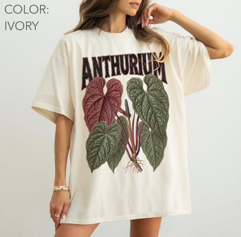May include: An ivory short-sleeved t-shirt featuring a graphic print of an Anthurium plant with large, textured green and deep red leaves. The word ANTHURIUM is printed in a bold, dark, distressed font above the botanical illustration.