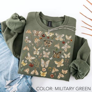 May include: A military green crewneck sweatshirt featuring a detailed graphic print of various moths in shades of brown, beige, and cream, interspersed with small botanical elements and flowers. A silver chain necklace rests on the neckline.