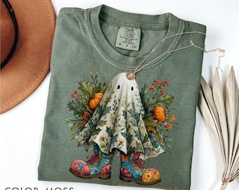May include: Moss green t-shirt featuring a whimsical ghost design. The ghost is adorned with floral details, pumpkins, and colorful boots. A silver necklace with a pendant rests on the shirt. The shirt is labeled "Comfort Colors".